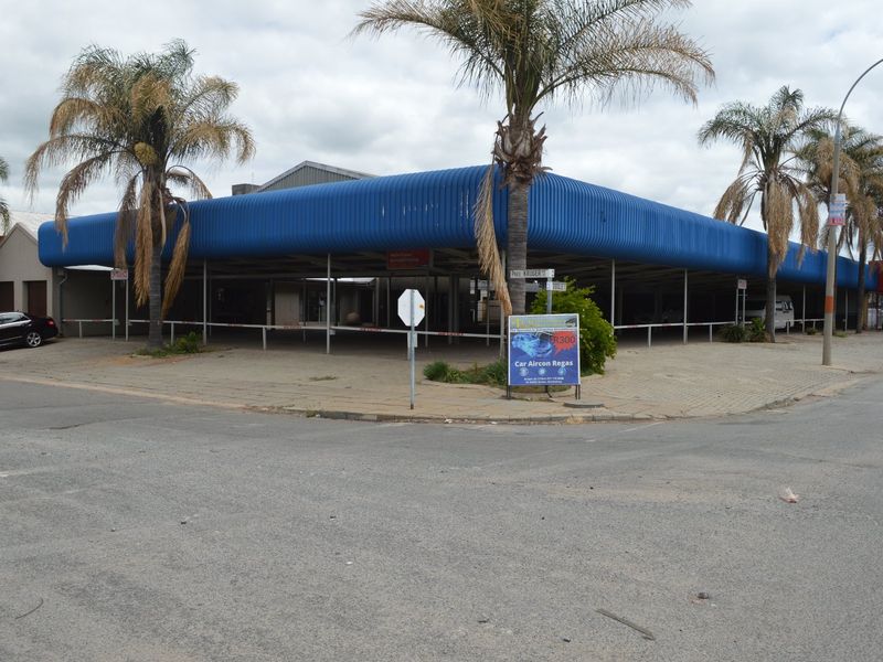 Corner commercial property featuring a large open-plan forecourt area with a prominent blue corrugated metal roof structure and paved exterior.