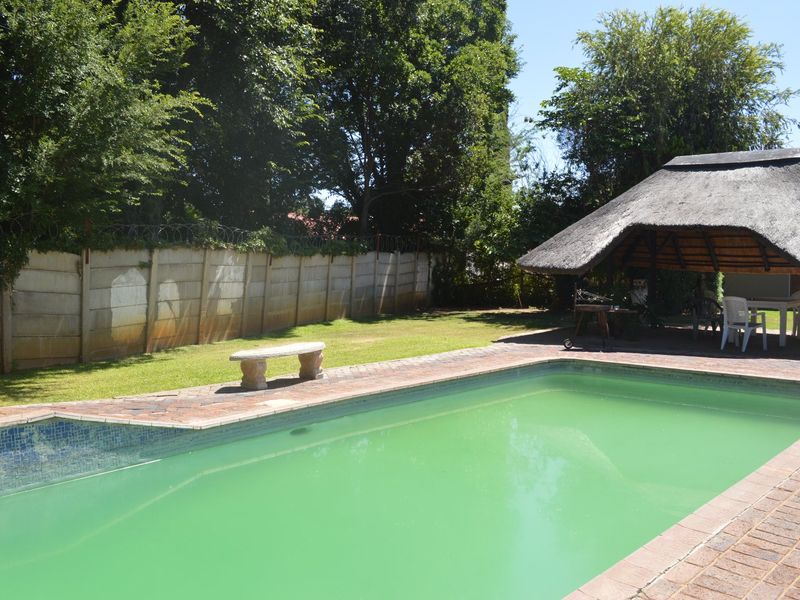 Outdoor pool area featuring a swimming pool, brick paving, and a thatched lapa structure set within a garden with perimeter walling.