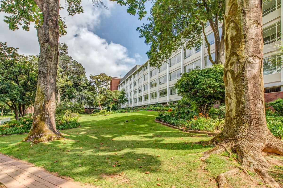 A view of the apartment building exterior featuring a well-maintained landscaped garden, mature trees, and a paved walkway.