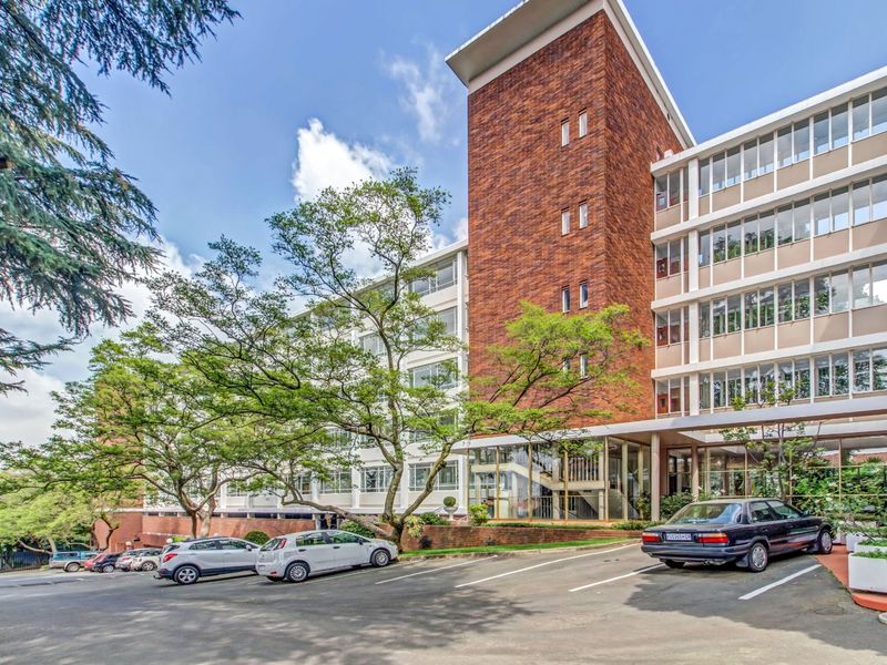 Exterior view of a multi-story apartment building featuring a brick facade, large windows, and a paved parking area at the front entrance.