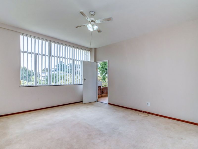 An empty bedroom featuring carpet flooring, a ceiling fan, and a large window with vertical blinds, offering access to an outdoor balcony area.