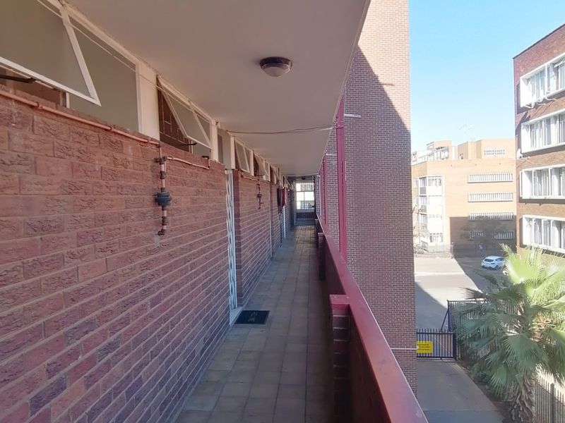 An exterior apartment balcony featuring exposed brick walls, tiled flooring, and a metal safety railing overlooking the complex grounds.