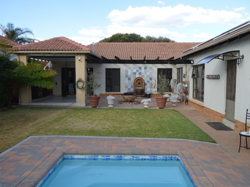 Rear exterior view featuring a swimming pool, paved patio area, covered veranda, and a landscaped garden with a decorative water feature.