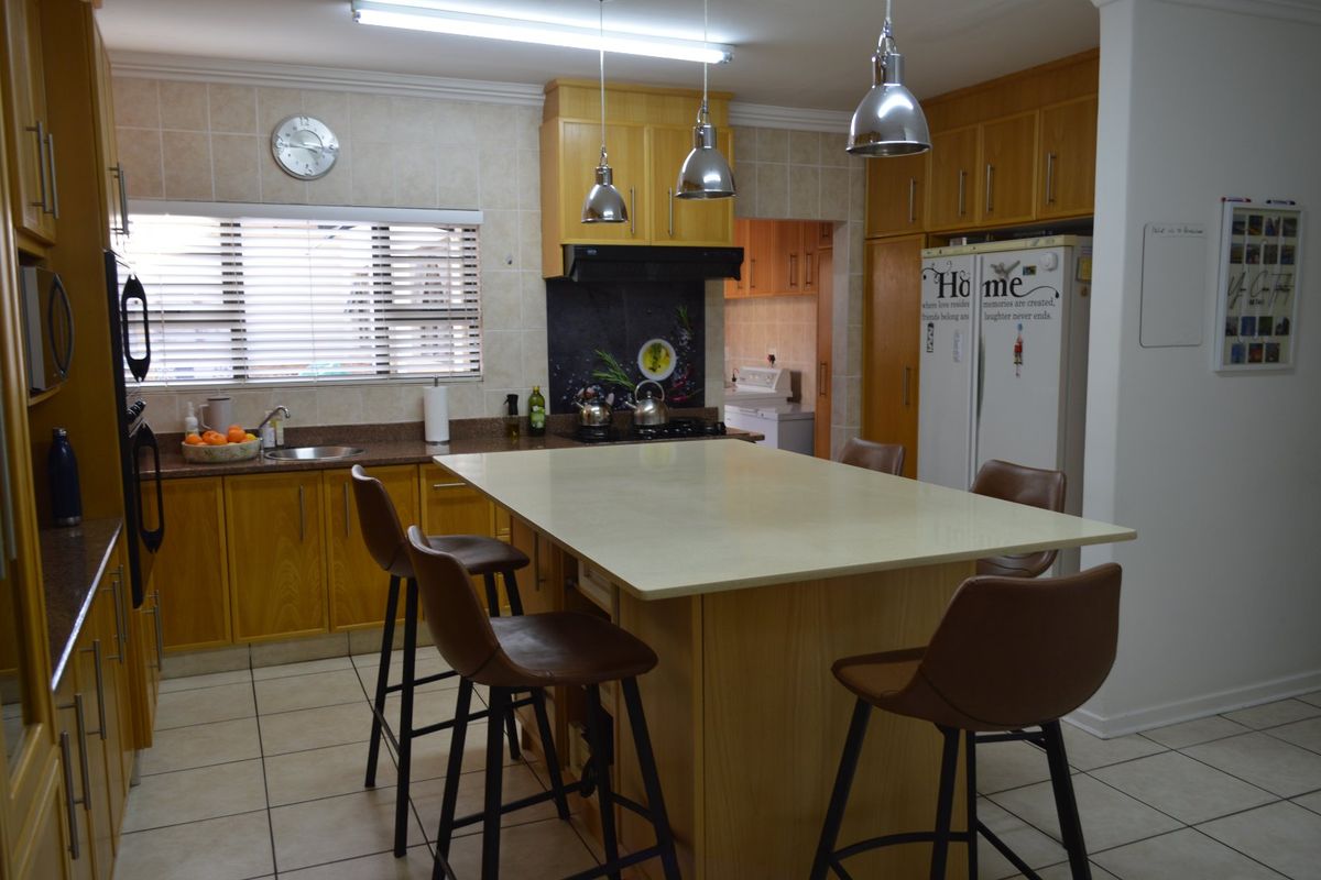 A spacious kitchen featuring wooden cabinetry, a central island with seating, stone countertops, and tiled flooring, with an adjacent scullery area.