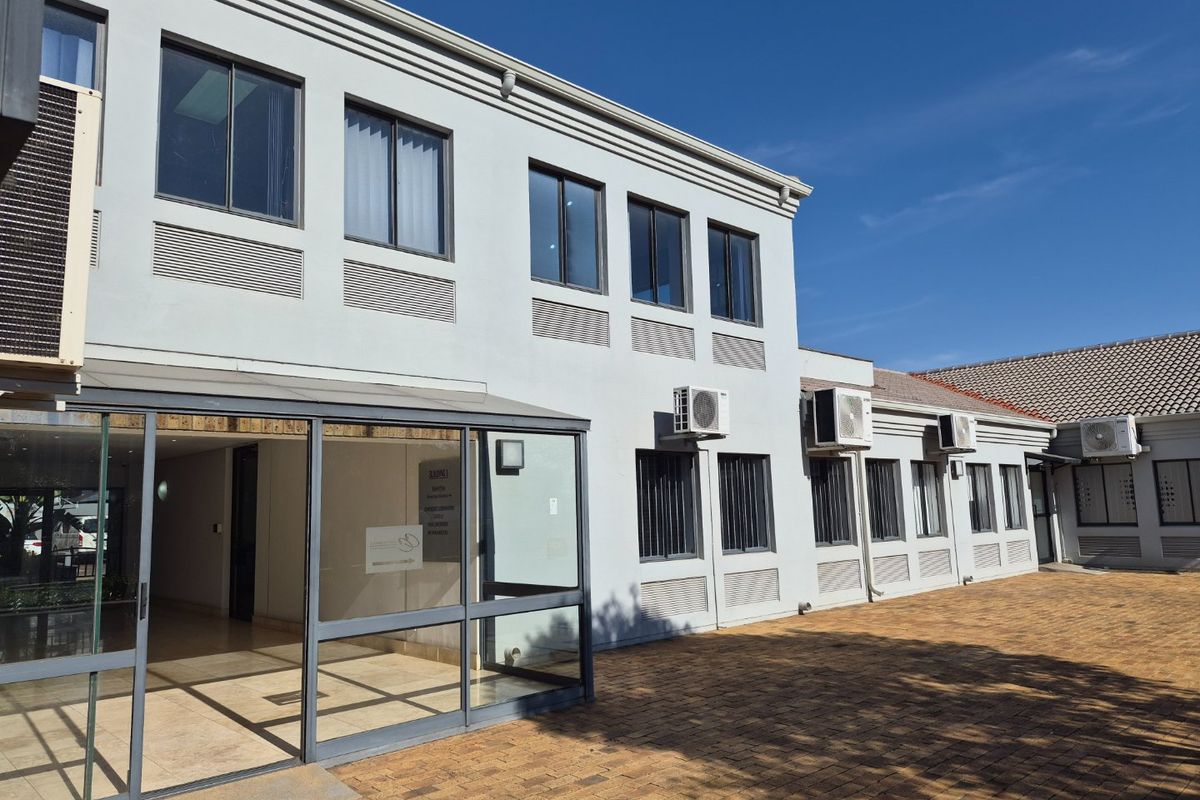 Rear exterior view of a commercial building featuring a paved courtyard, glass-enclosed entrance, and multiple wall-mounted air conditioning units.