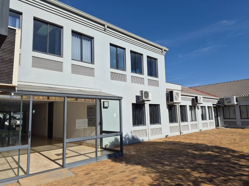 Rear exterior view of a commercial building featuring a paved courtyard, glass-enclosed entrance, and multiple wall-mounted air conditioning units.