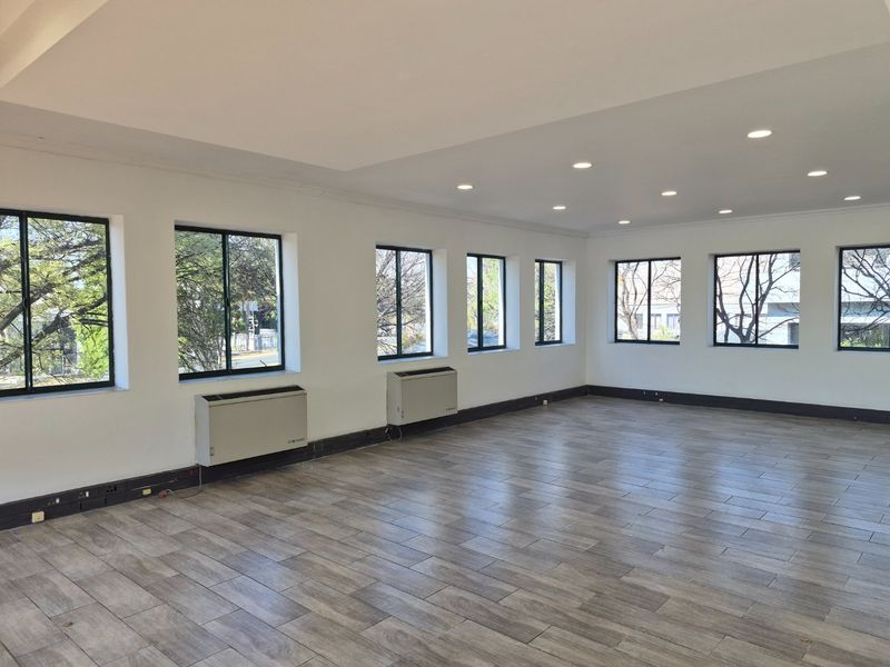A bright, vacant commercial office space featuring wood-look tiled flooring, recessed ceiling lighting, and multiple windows providing natural light.
