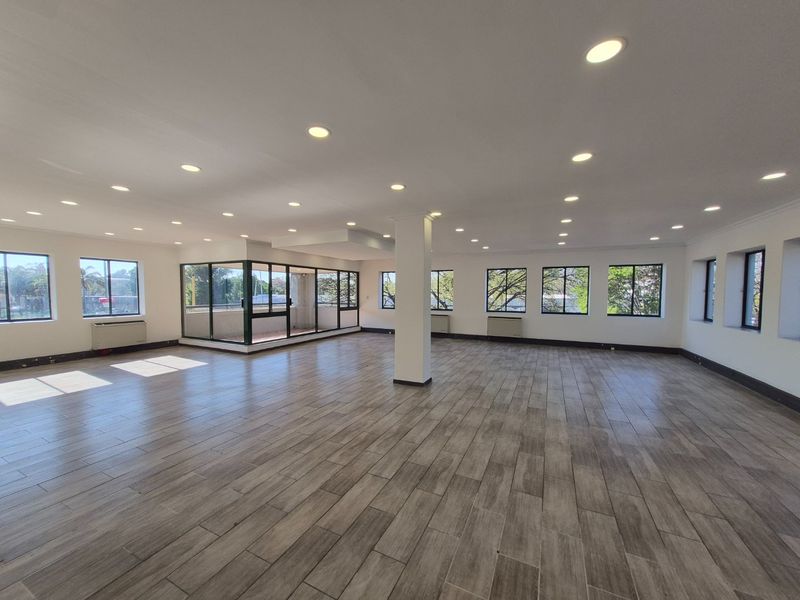 Spacious, open-plan commercial office area featuring wood-look tile flooring, recessed lighting, and large windows providing ample natural light.