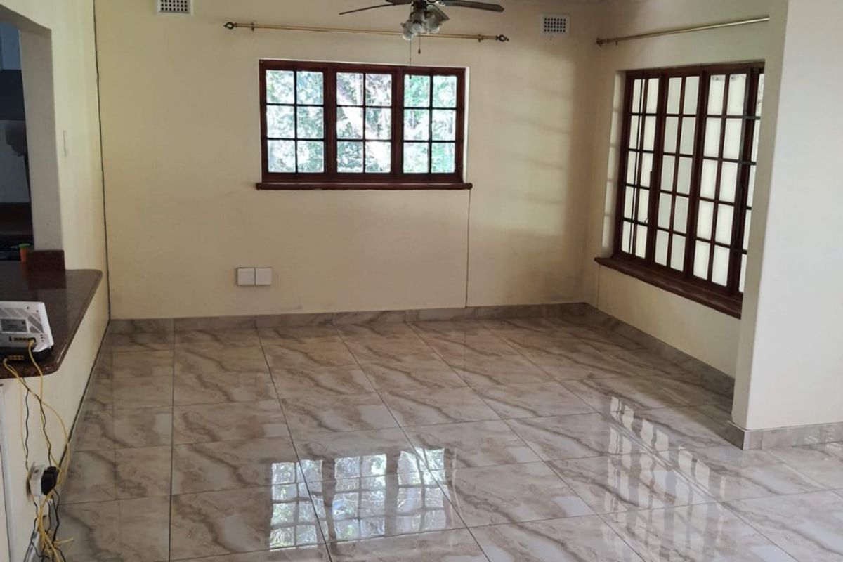 A bright, empty living area featuring polished tiled flooring, decorative ceiling cornicing, a ceiling fan, and large wooden-framed windows.