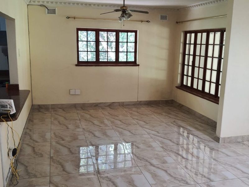 A bright, empty living area featuring polished tiled flooring, decorative ceiling cornicing, a ceiling fan, and large wooden-framed windows.
