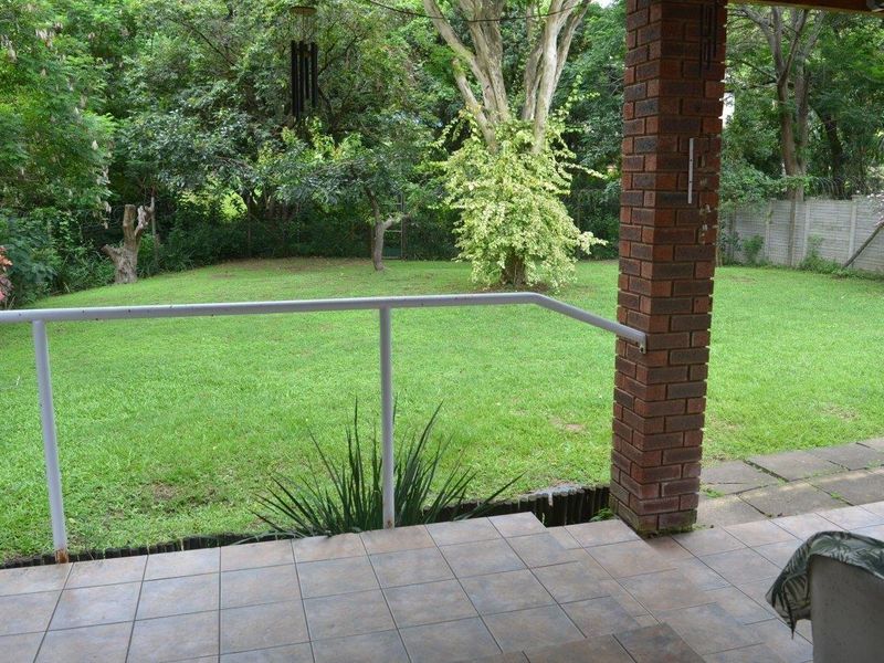 A covered patio area featuring tiled flooring and a brick pillar, overlooking a spacious private garden with mature trees and perimeter fencing.