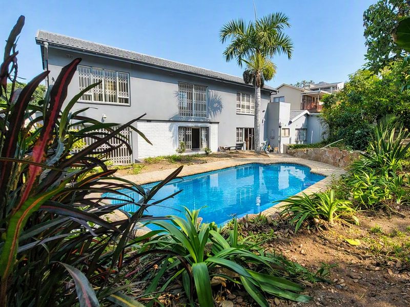 Rear view of a two-story house featuring a swimming pool, paved surround, and stone retaining wall set within a landscaped garden.