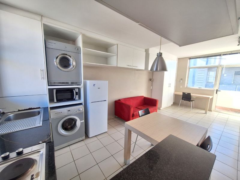 Open-plan apartment living area featuring a compact kitchen with built-in appliances, tiled flooring, and a dedicated study nook near the balcony entrance.