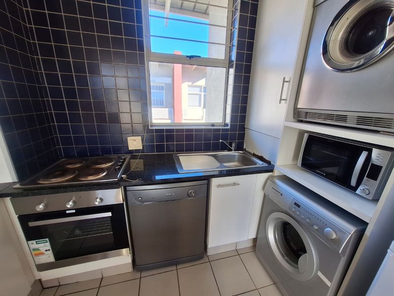 Compact kitchen featuring built-in oven, electric hob, stainless steel sink, granite countertops, and dark tiled backsplash with integrated laundry appliance space.