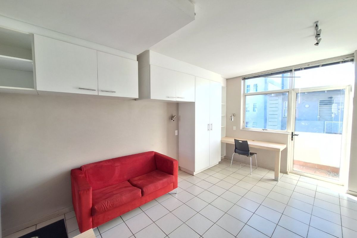 A functional bedroom featuring built-in cupboards, a dedicated desk area, tiled flooring, and direct access to an outdoor balcony through a glass door.