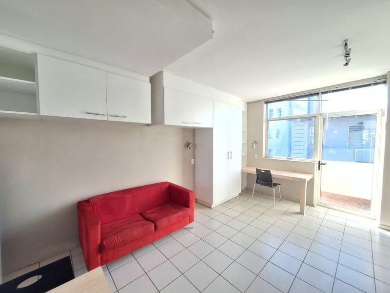 A functional bedroom featuring built-in cupboards, a dedicated desk area, tiled flooring, and direct access to an outdoor balcony through a glass door.