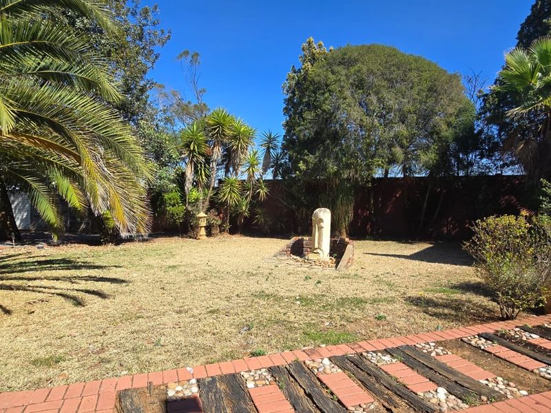 A spacious garden area featuring a brick-paved pathway, railway sleeper accents, a central stone water feature, and established trees against a brick building facade.