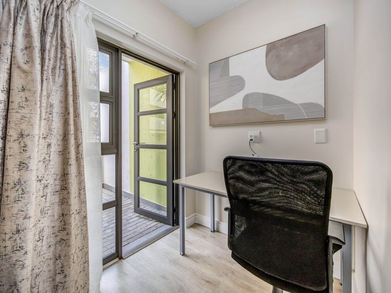 A bright home office space featuring laminate flooring and direct access to a small outdoor patio area through an aluminium framed glass door.