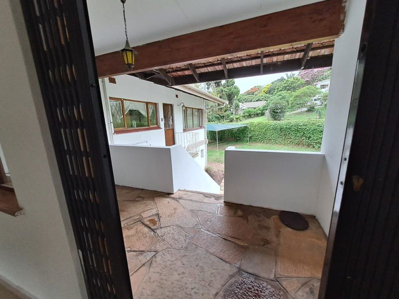 A covered patio area featuring rustic flagstone flooring, a timber beam ceiling, and a wrought iron railing overlooking the rear garden.