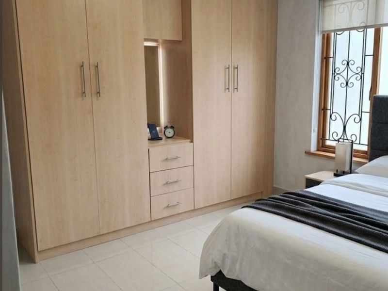 A well-maintained bedroom featuring light wood built-in cupboards, tiled flooring, and a window fitted with security bars and a roller blind.