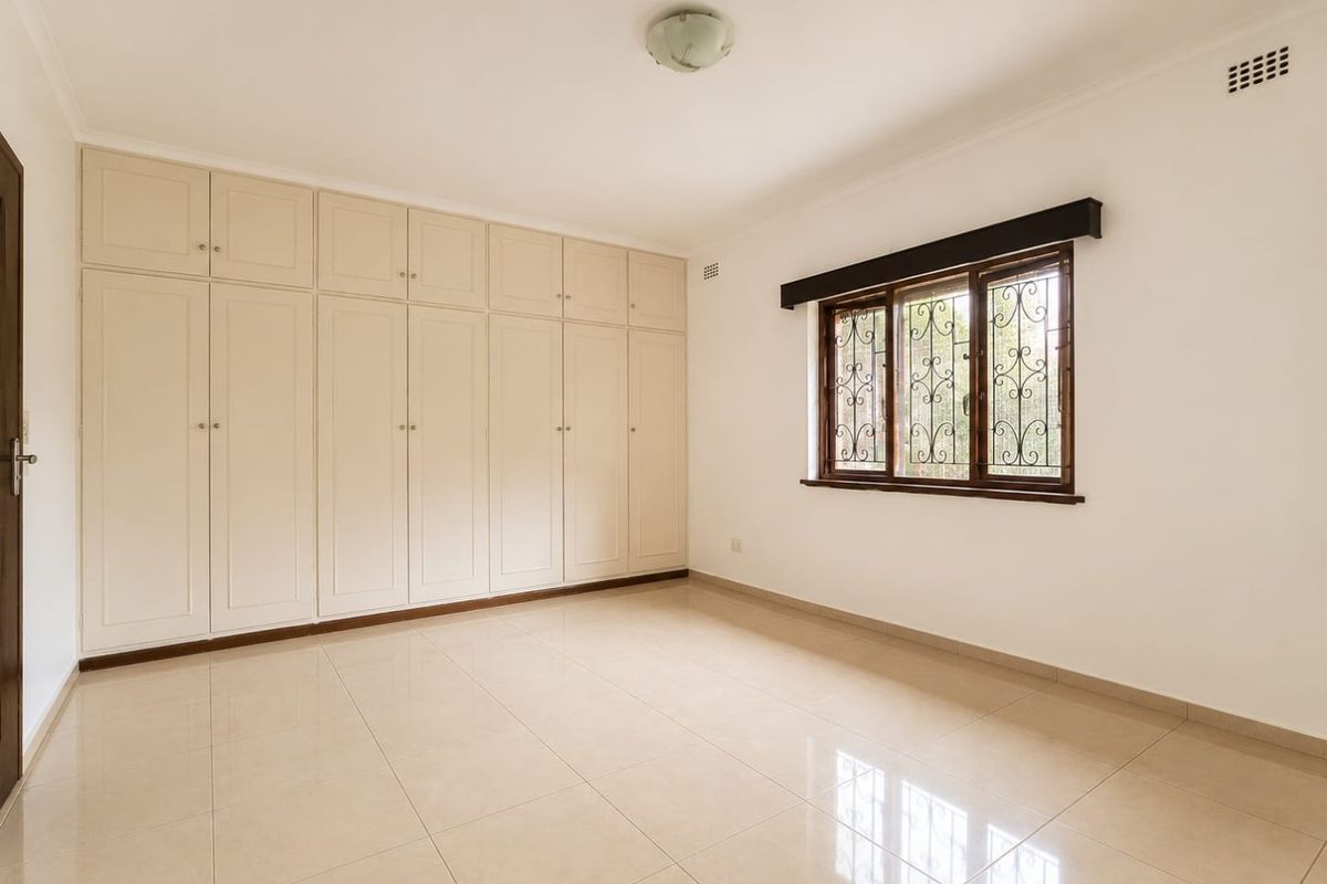 A bright, empty bedroom featuring neutral tiled flooring and extensive built-in storage cupboards with a window fitted with security bars.