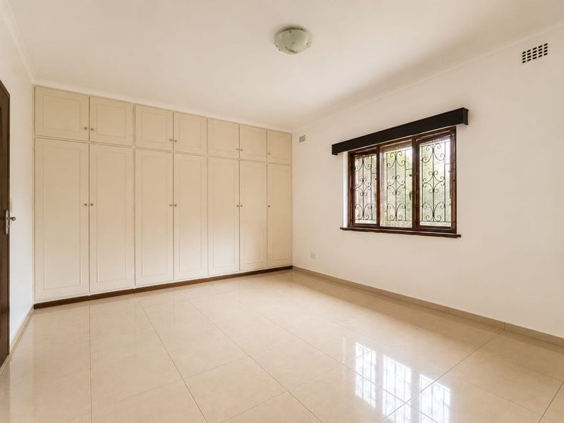 A bright, empty bedroom featuring neutral tiled flooring and extensive built-in storage cupboards with a window fitted with security bars.