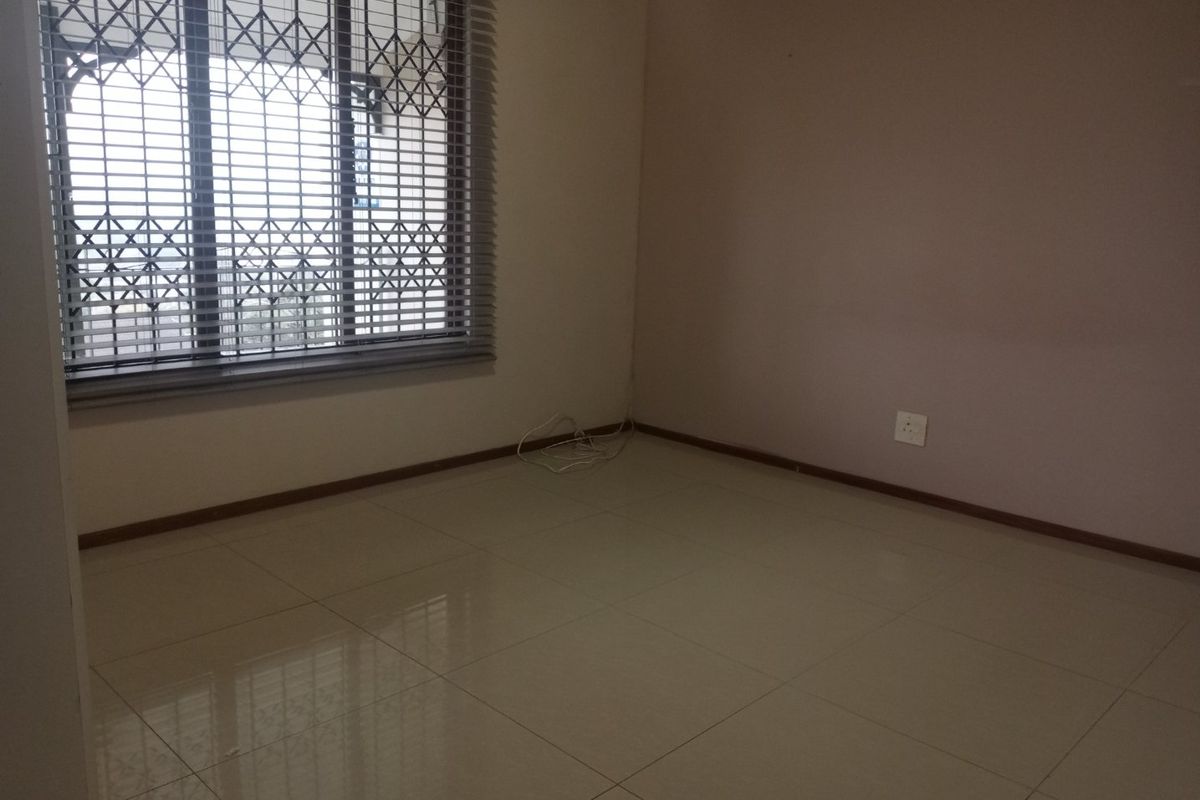 An empty bedroom featuring neutral tiled flooring, a window fitted with security bars and venetian blinds, and standard electrical fixtures.