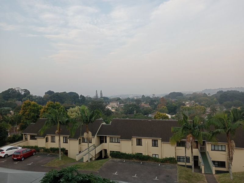 An elevated aerial view of an apartment complex featuring tiled roofs, paved parking areas, and established garden landscaping with palm trees.