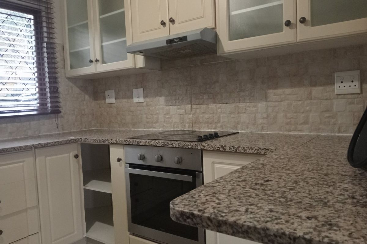 A functional kitchen featuring granite countertops, built-in oven, electric hob, extractor fan, and ample shaker-style cabinetry with a tiled backsplash.