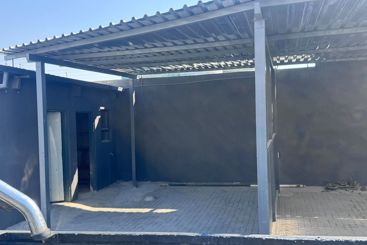 A covered outdoor area featuring a corrugated metal roof supported by a steel frame, with paved flooring and enclosed by a boundary wall.