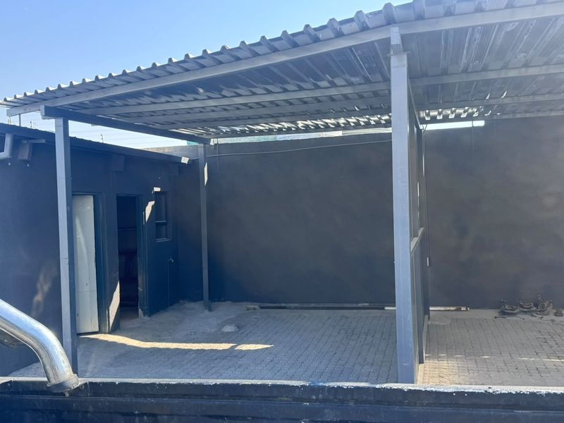 A covered outdoor area featuring a corrugated metal roof supported by a steel frame, with paved flooring and enclosed by a boundary wall.