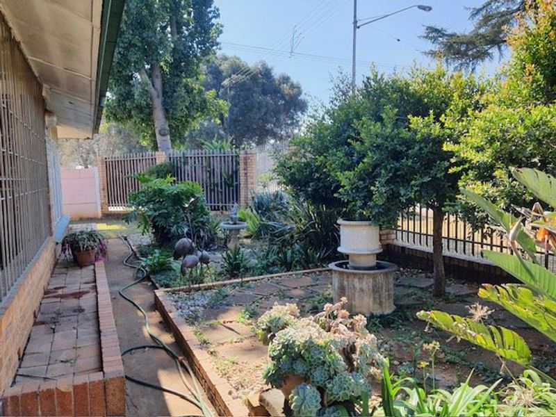 A residential garden area featuring brick paving, established landscaping, a stone fountain base, and metal perimeter fencing.
