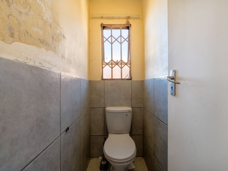 A small, functional guest toilet featuring tiled walls and a security gate on the window, currently requiring cosmetic wall repairs and maintenance.