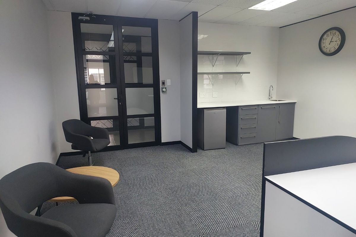A modern commercial office kitchenette featuring grey cabinetry, a sink, floating shelves, and a glass-panelled entrance door with carpeted flooring.