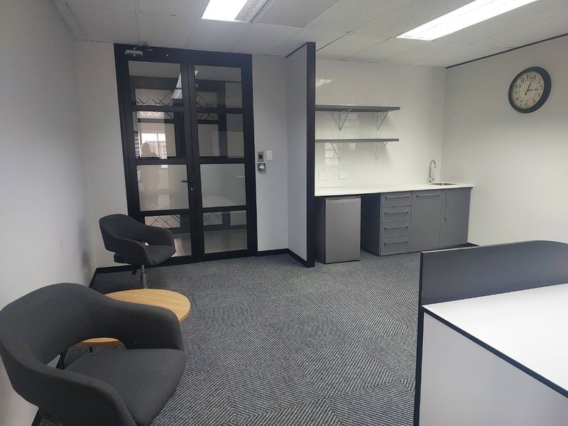 A modern commercial office kitchenette featuring grey cabinetry, a sink, floating shelves, and a glass-panelled entrance door with carpeted flooring.