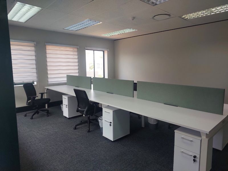A professional office space featuring built-in desk workstations with privacy partitions, carpeted flooring, recessed ceiling lighting, and adjustable window blinds.