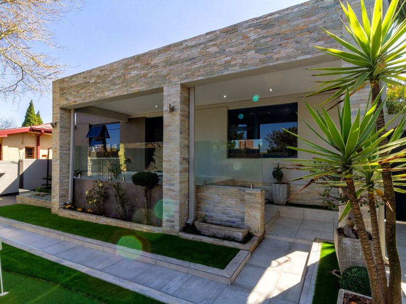 A modern covered patio featuring stone cladding, glass balustrades, and recessed lighting, overlooking a well-maintained garden area.