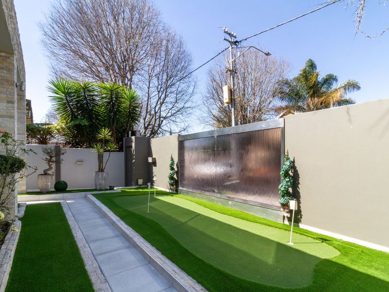 A well-maintained private garden featuring a professional-style artificial turf putting green, paved walkway, and a modern wall-mounted water feature.