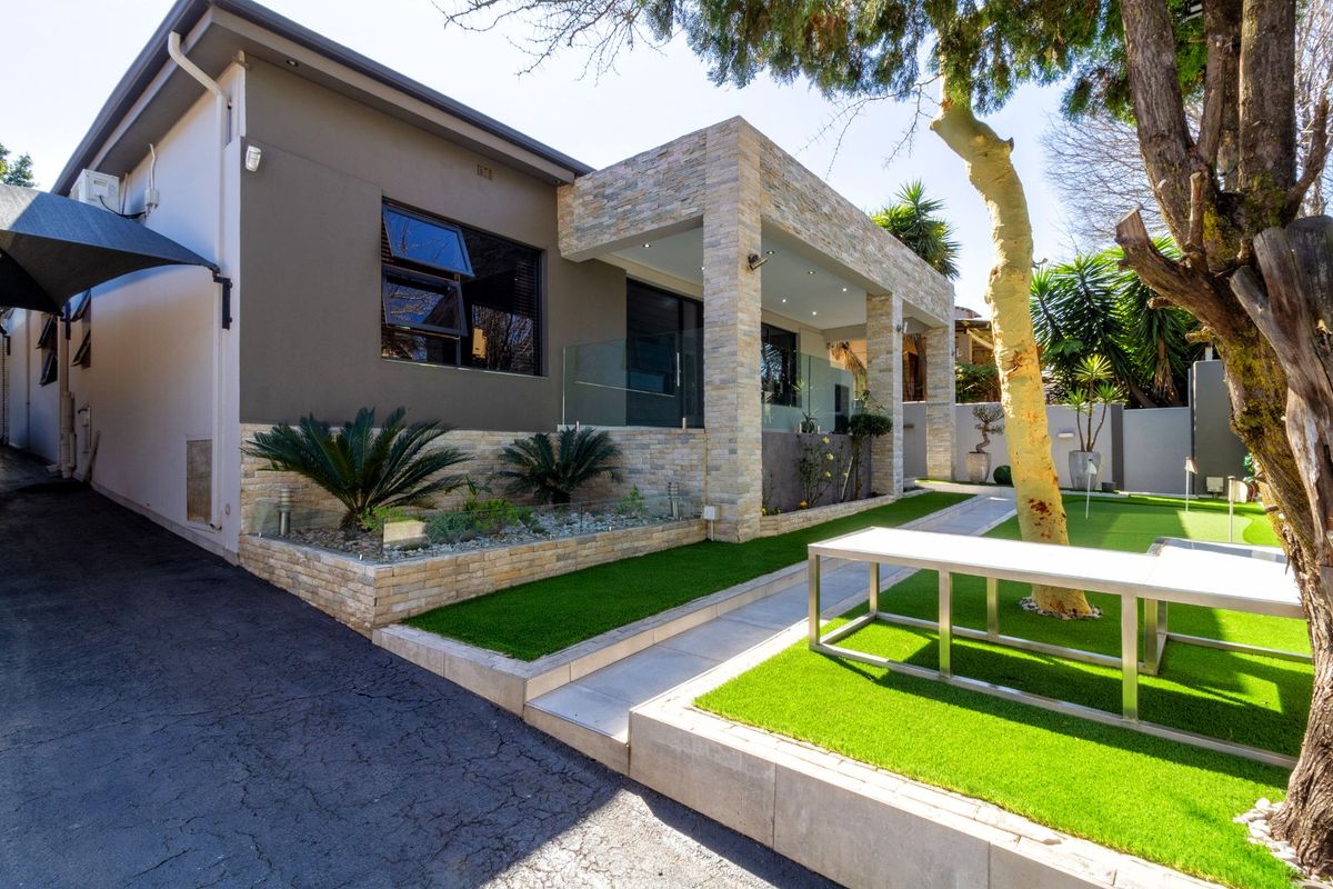 Modern rear exterior featuring stone-clad pillars, a glass balustrade, artificial lawn, and a paved walkway leading to a covered patio area.