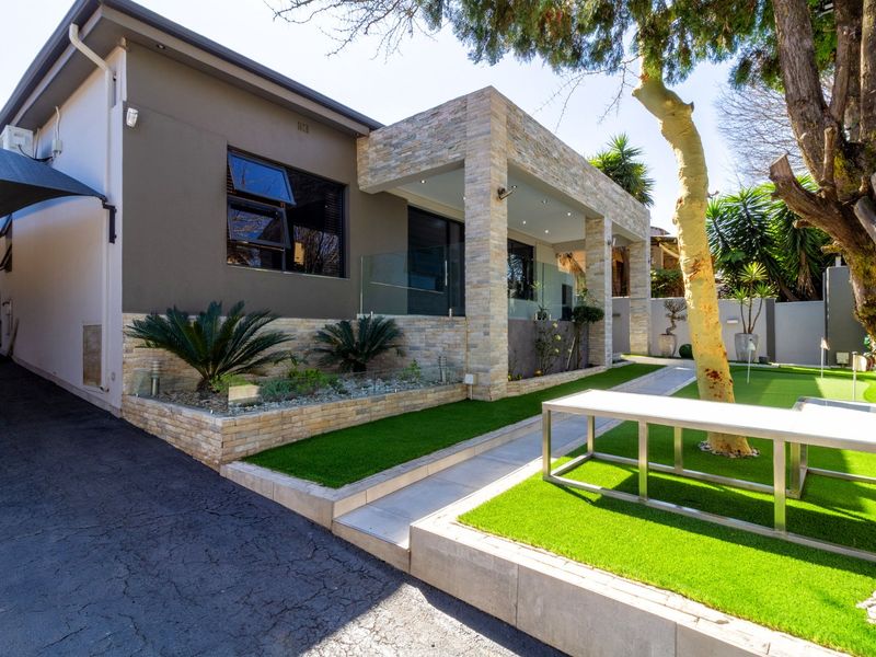 Modern rear exterior featuring stone-clad pillars, a glass balustrade, artificial lawn, and a paved walkway leading to a covered patio area.