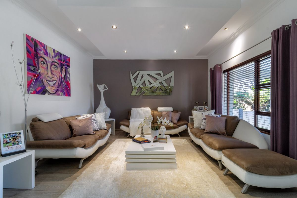 A well-maintained living room featuring a tray ceiling with recessed downlights, laminate flooring, and large windows with venetian blinds.