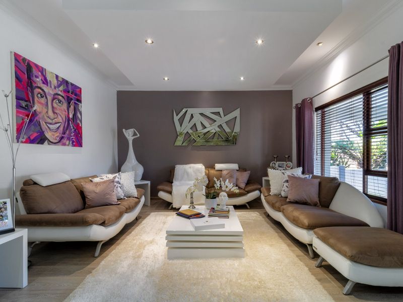 A well-maintained living room featuring a tray ceiling with recessed downlights, laminate flooring, and large windows with venetian blinds.