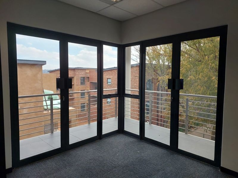 Empty office space featuring large black-framed glass doors opening onto a corner balcony with stainless steel balustrades and views of the surrounding complex.