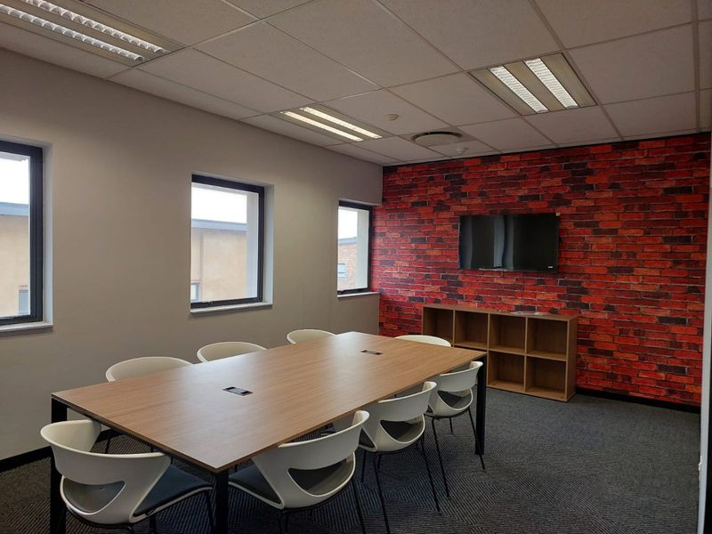 A modern office meeting room featuring a brick-effect accent wall, suspended ceiling with recessed lighting, and carpeted flooring.