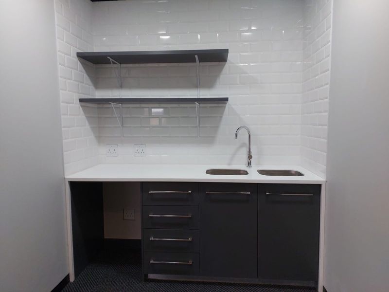 A clean, modern kitchenette or scullery area featuring dark grey cabinetry, white subway tile backsplash, stainless steel sink, and floating shelving.