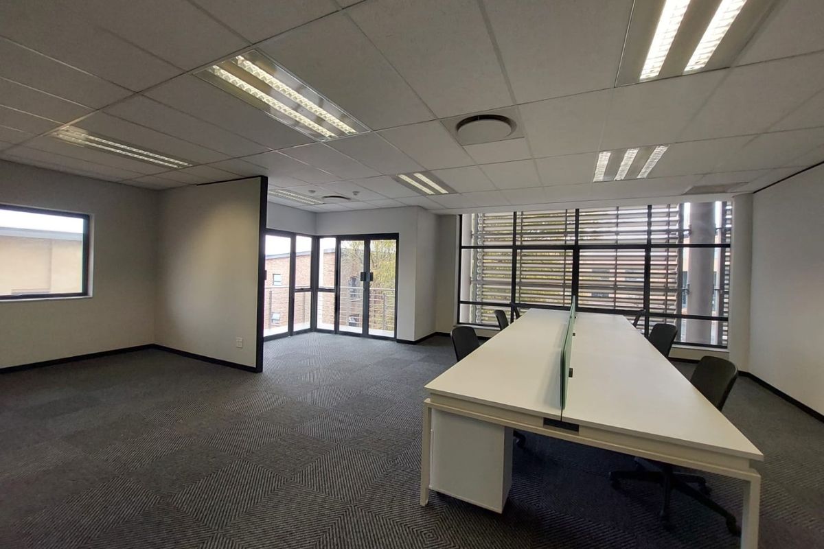 A bright, modern office space featuring carpeted flooring, suspended ceilings with recessed lighting, and large windows with external louvers for natural light control.