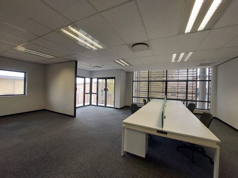 A bright, modern office space featuring carpeted flooring, suspended ceilings with recessed lighting, and large windows with external louvers for natural light control.