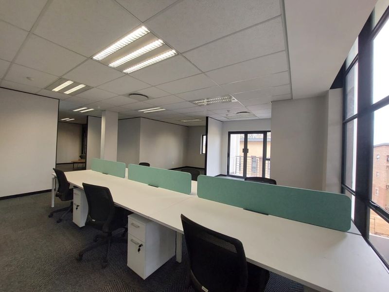 Modern open-plan office space featuring suspended ceilings, recessed lighting, carpeted flooring, and large windows providing ample natural light and balcony access.