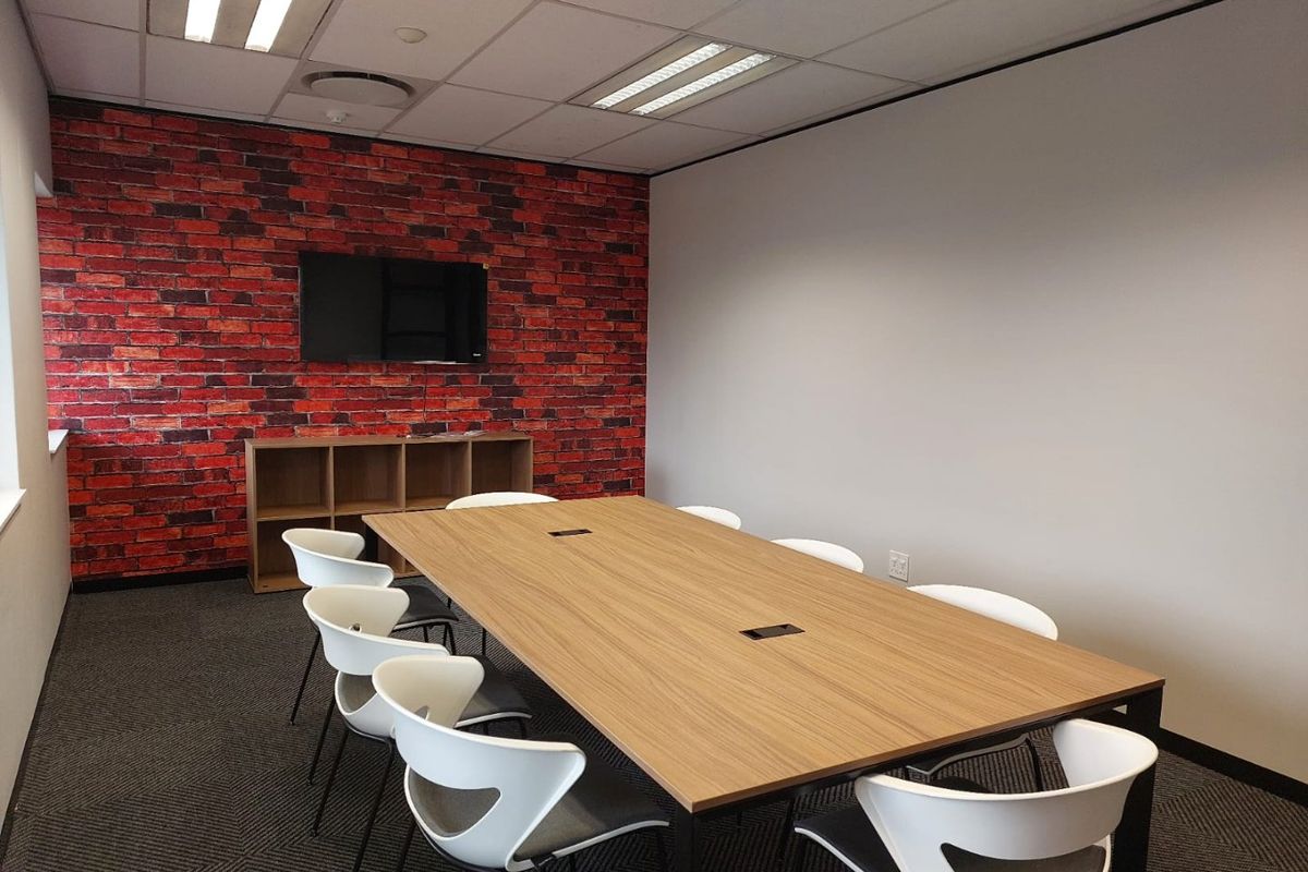 A professional office meeting room featuring a modern boardroom table, suspended ceiling, and a distinctive brick-effect feature wall.
