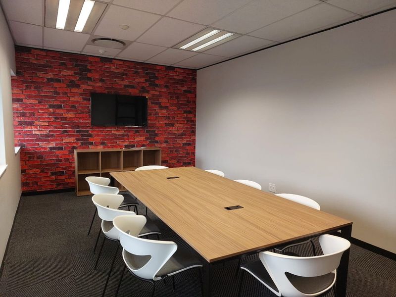 A professional office meeting room featuring a modern boardroom table, suspended ceiling, and a distinctive brick-effect feature wall.